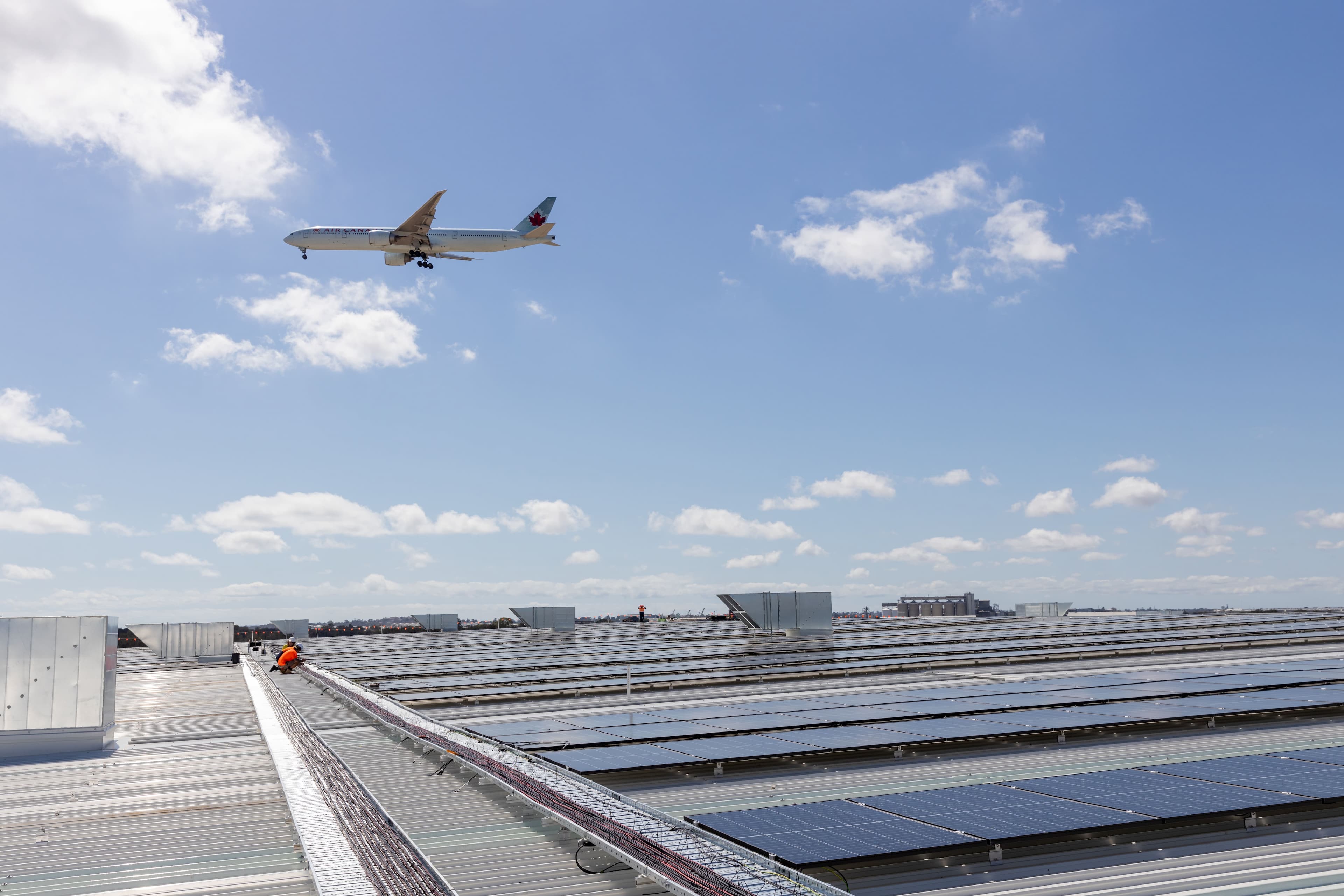 A commercial airplane flies over a roof with installed solar panels under a clear sky. Two workers in orange uniforms are visible on the rooftop.