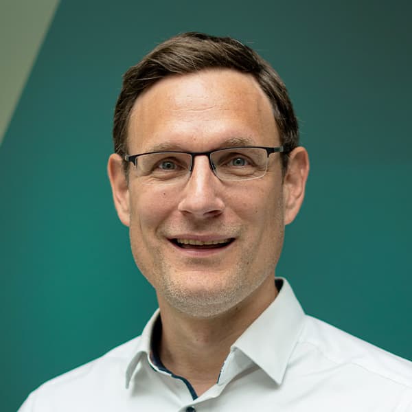 A man with short brown hair, wearing glasses and a white collared shirt, smiles in front of a green background.