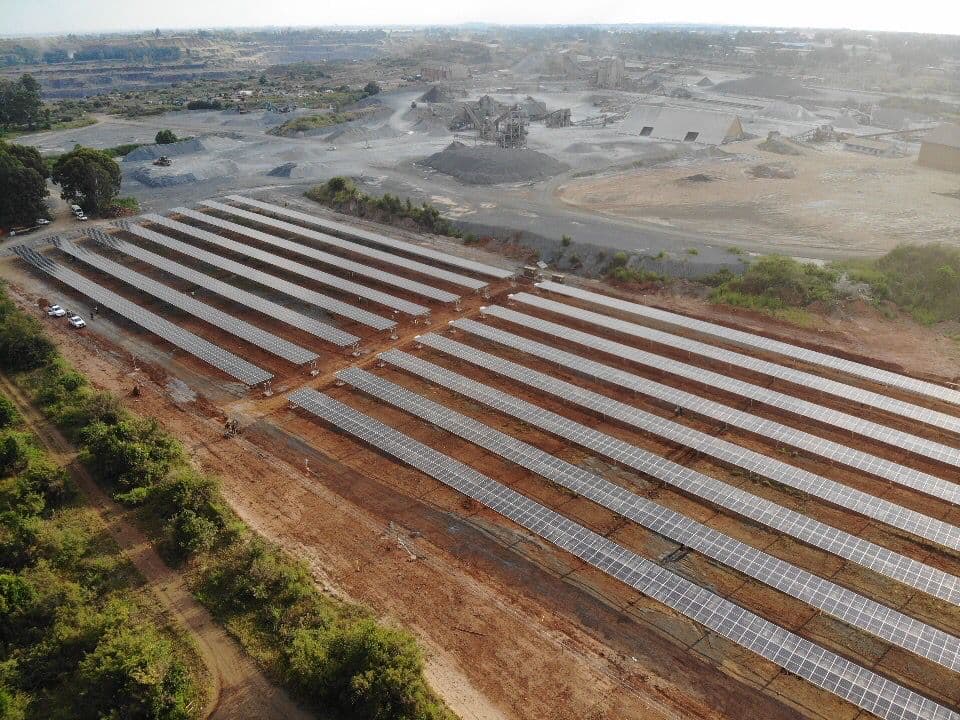 Aerial view of a large solar farm with multiple rows of solar panels installed on a vast, barren land, with industrial buildings and sparse vegetation in the background.