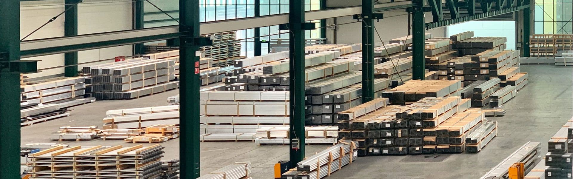 A large industrial warehouse with stacks of organized materials, including metal beams and sheets, on the floor and shelves. The warehouse is spacious with high ceilings and green metal pillars.