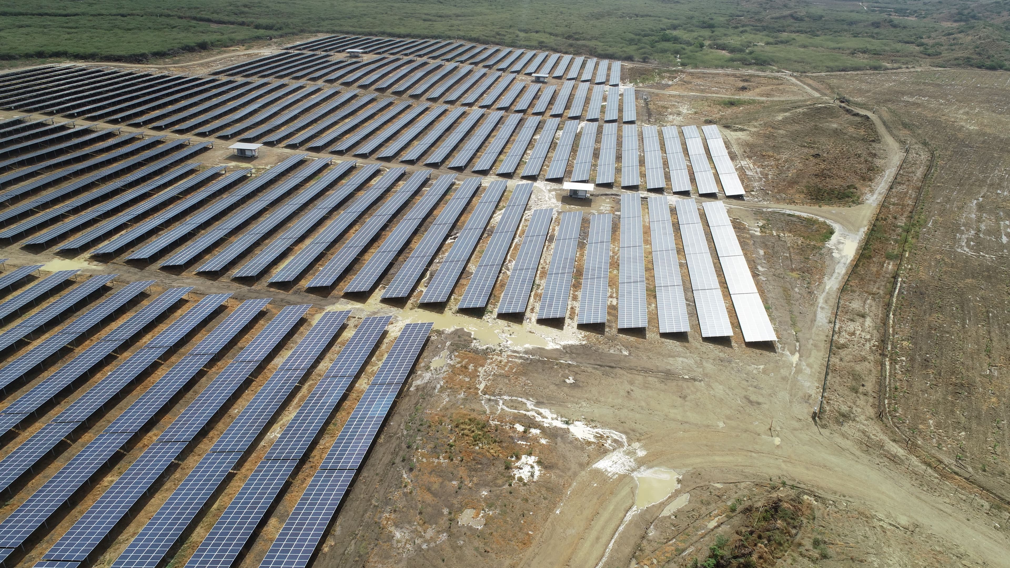 Aerial view of a large solar farm with rows of solar panels set up on a dry, open landscape. Dirt roads and patches of greenery can be seen around the installation.
