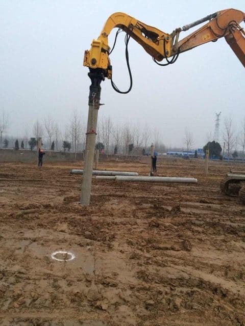 A piece of heavy machinery drives a concrete pile into the ground at a construction site. Several workers stand in the background on the muddy site.