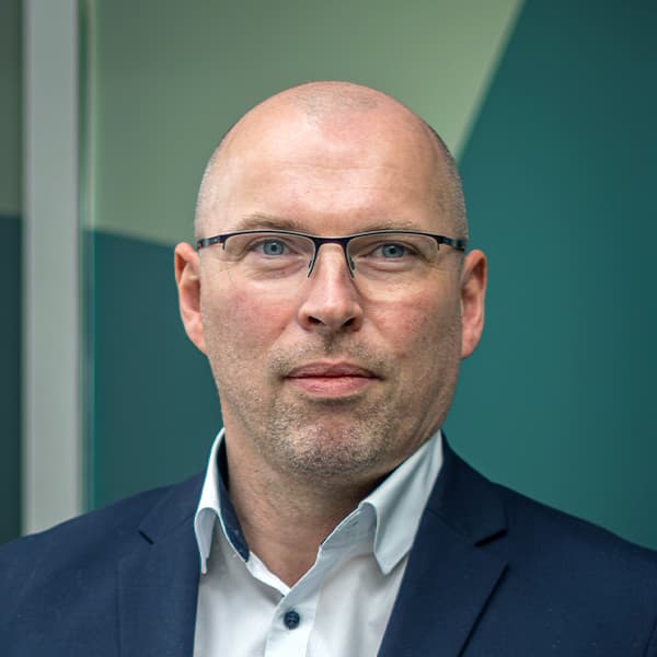 A middle-aged man wearing glasses and a dark blazer stands in front of a green geometric background, looking directly at the camera.