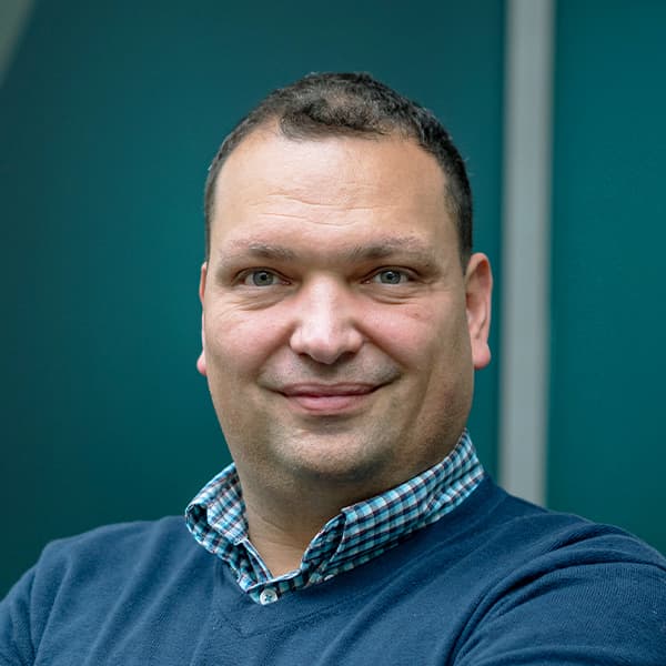 A person with short hair, wearing a checked shirt and a dark blue sweater, poses in front of a teal background.