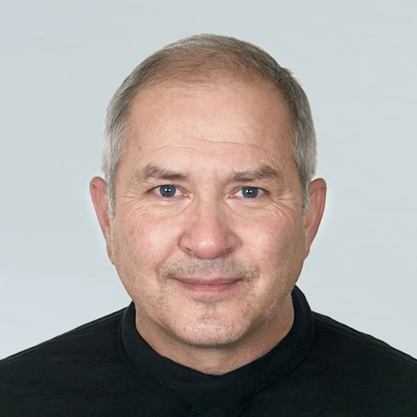 A man with short, graying hair and a neutral expression is wearing a black, high-collared shirt. The background is plain and light-colored.