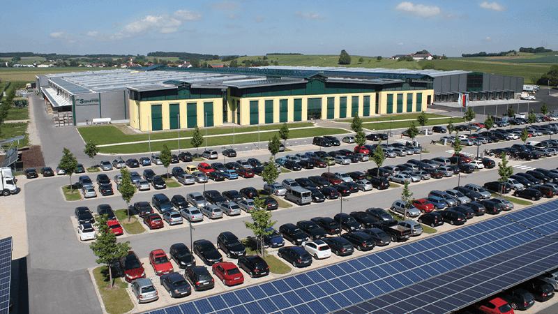 A modern industrial building with green and yellow facade is surrounded by a large parking lot with numerous cars. Solar panels are visible in the foreground, and a grassy landscape extends into the background.