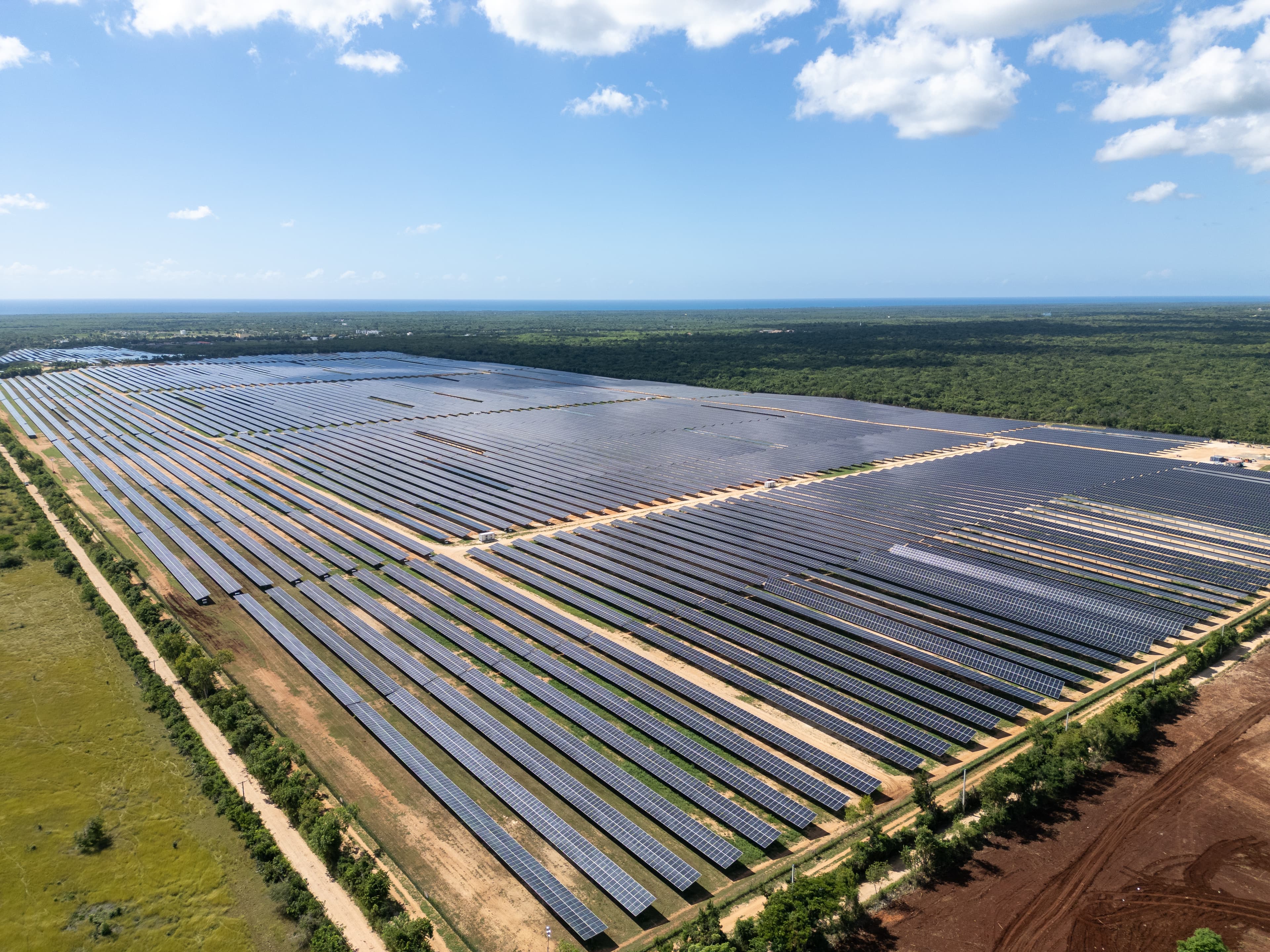 Aerial view of a large solar farm with numerous rows of solar panels set in a landscape of green fields and trees, under a partly cloudy sky.