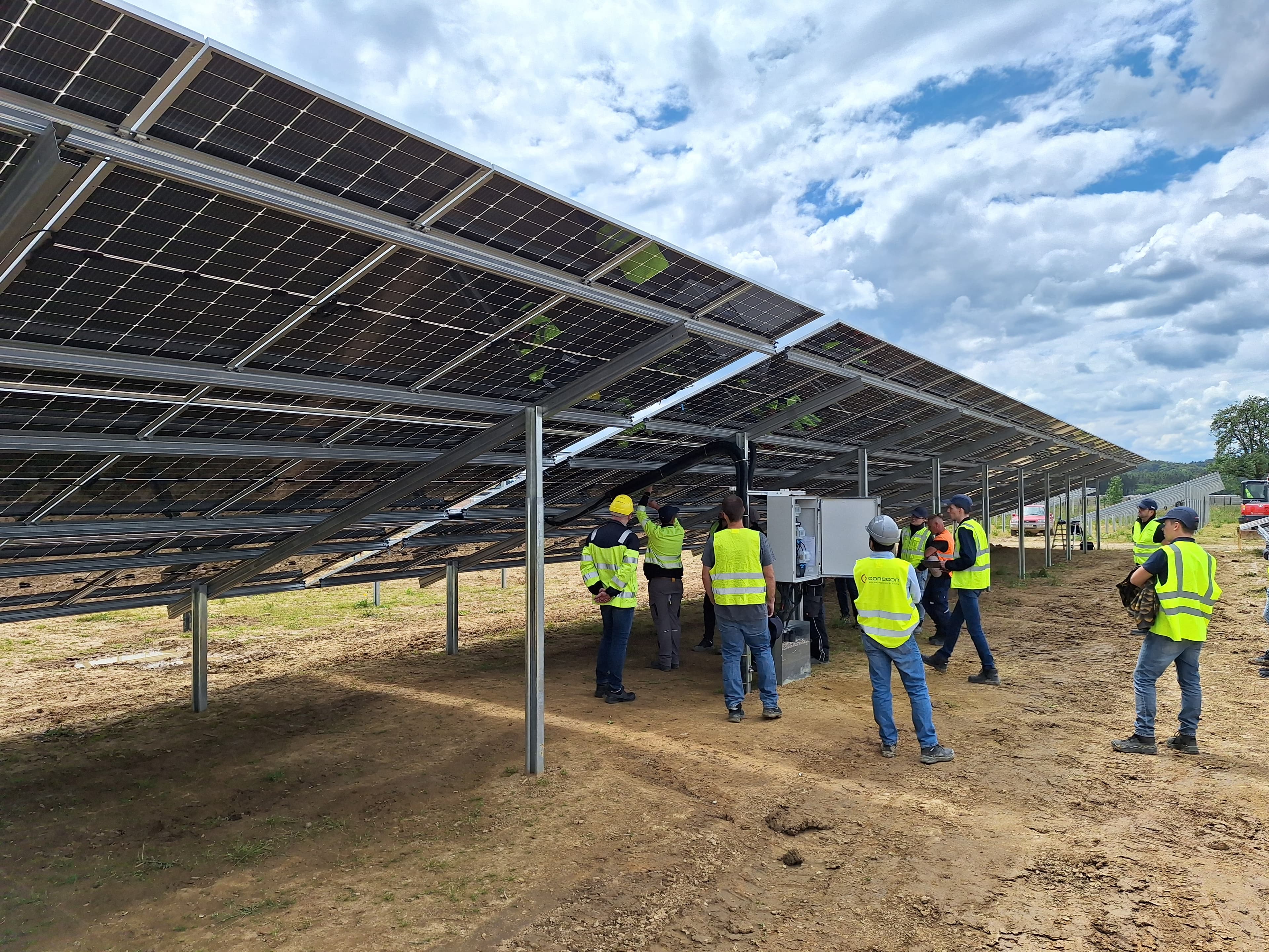 Workers in high-visibility vests and helmets install and inspect solar panels mounted on metal structures in an open construction site.