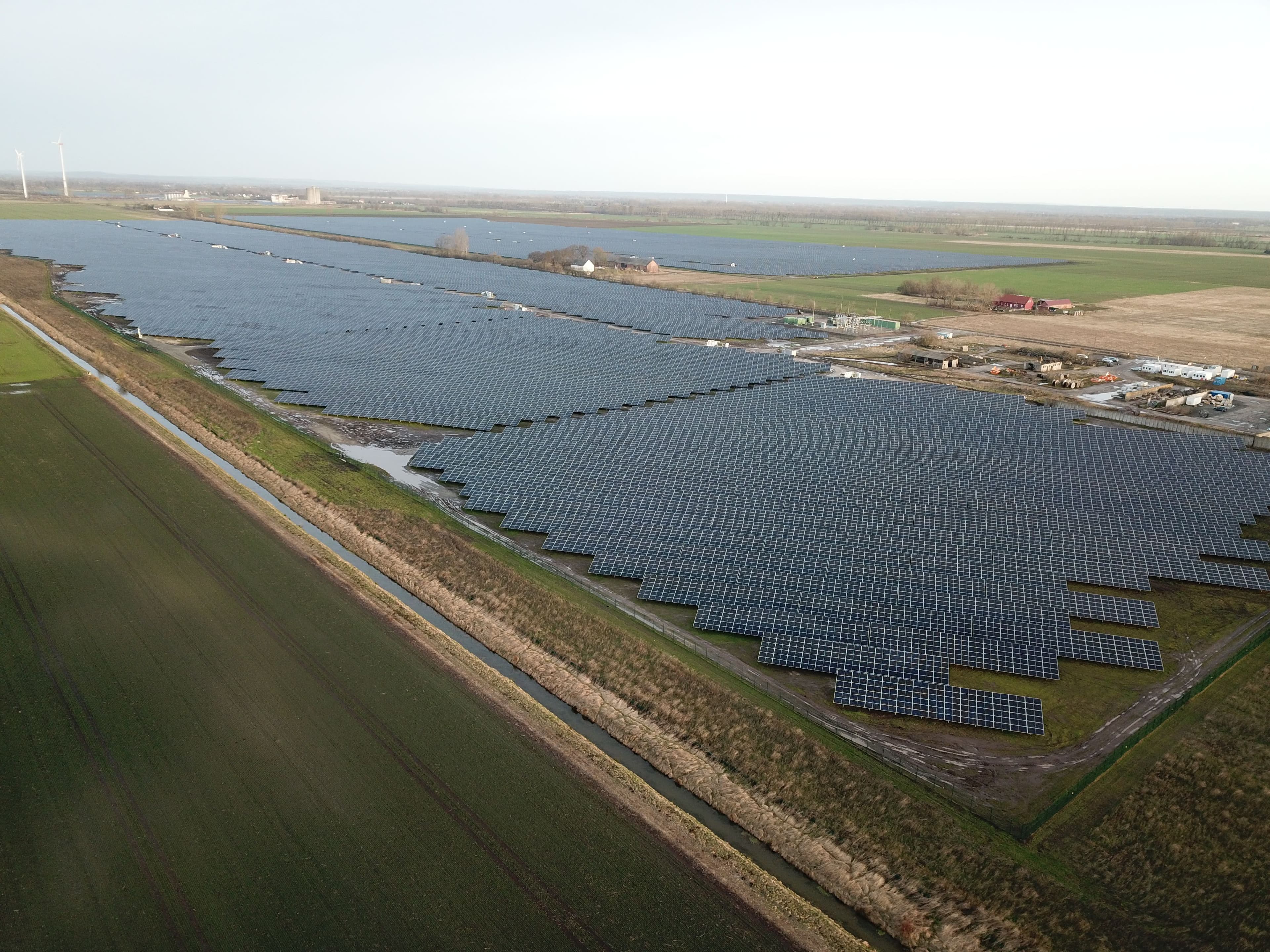 Aerial view of a large solar panel farm in a rural area, with surrounding greenery and distant buildings.