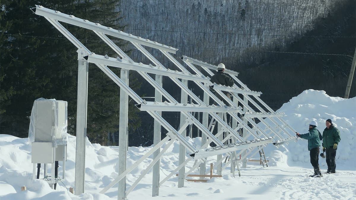 Workers install a metal frame for solar panels in a snowy area surrounded by trees and hills.