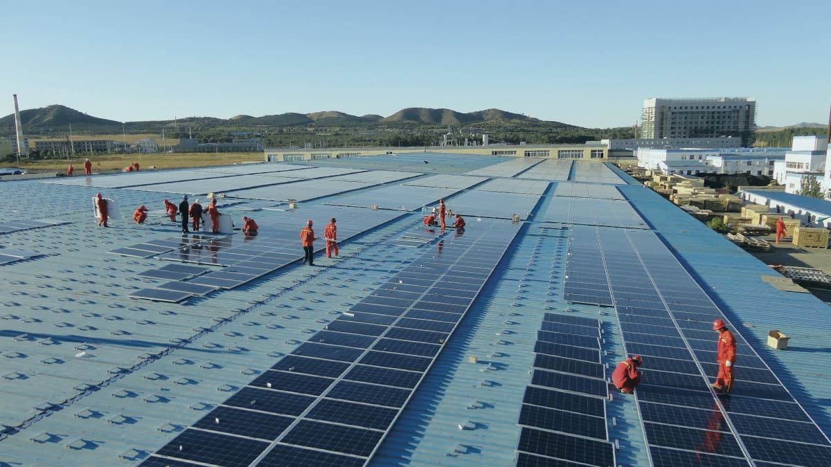 Workers in red uniforms are installing solar panels on a large, flat rooftop in an industrial area with hills in the background.