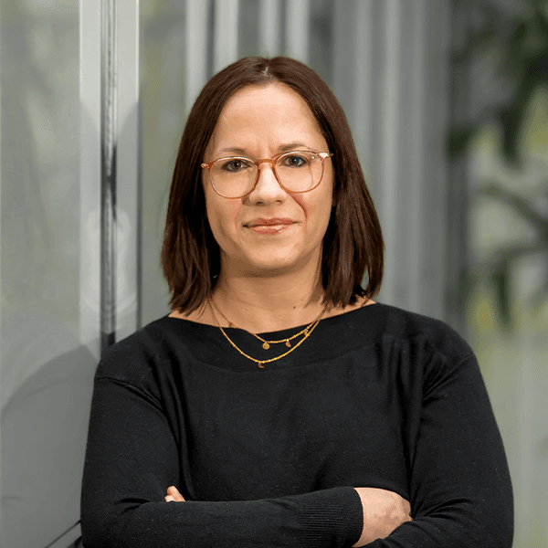 A person with short brown hair, wearing glasses and a black top, stands with folded arms in front of a reflective surface.