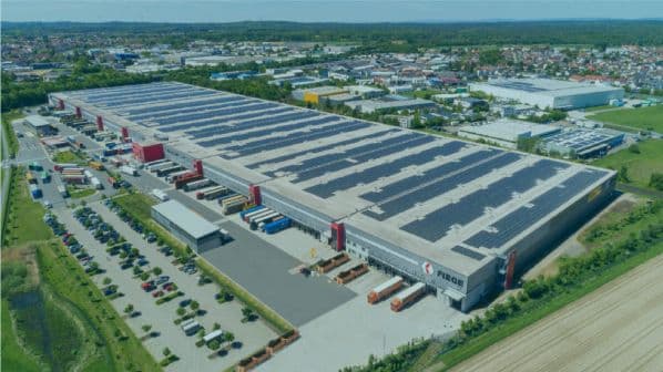 Aerial view of a large industrial warehouse complex with rows of solar panels on the roof, parking lots, and loading bays with trucks. Surrounding area includes other buildings and green spaces.