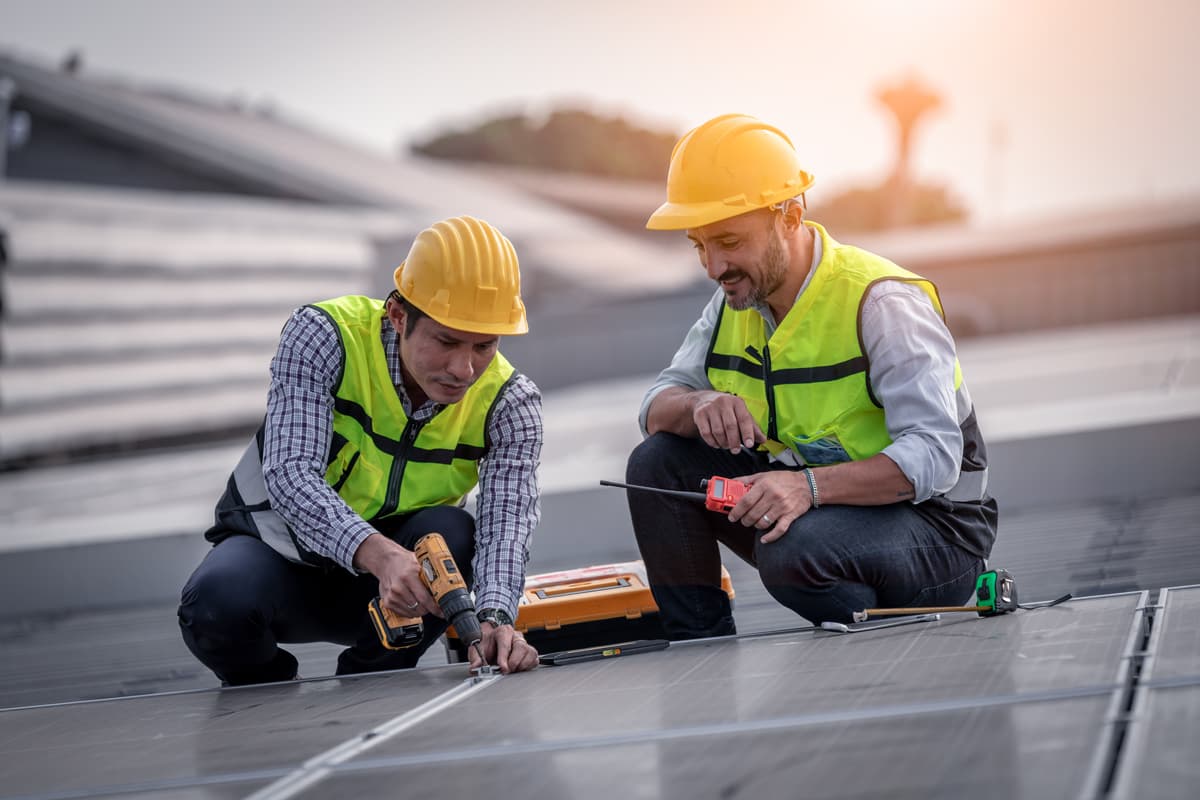 Two workers wearing safety gear install solar panels using tools on a rooftop.