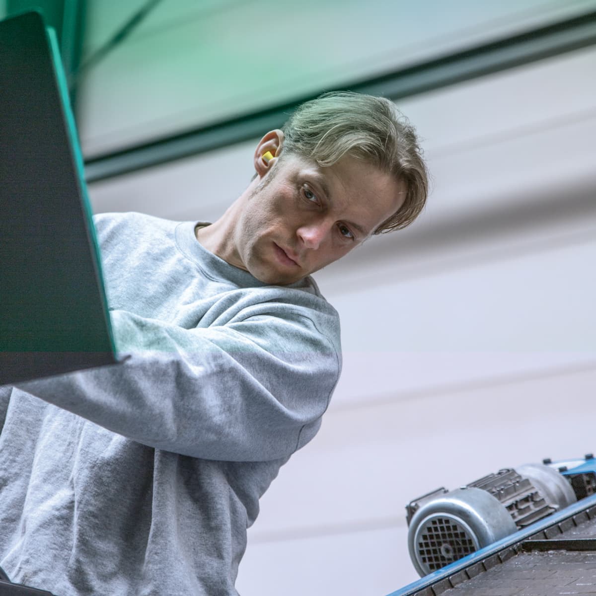 A person wearing a gray sweatshirt and ear protection operates machinery or equipment indoors.