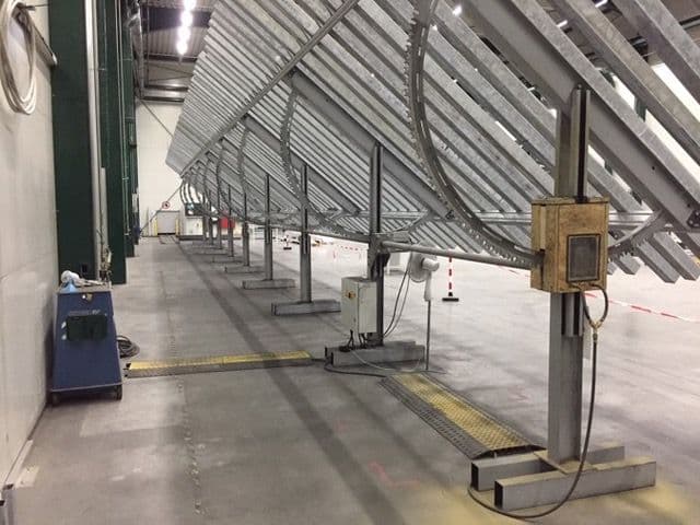 A large industrial machine with angled metal beams is installed in a spacious indoor facility with concrete flooring and high ceilings.