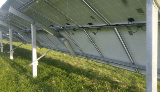 A row of solar panels installed on metal supports in a grassy field, shown from the rear perspective.