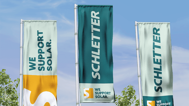 Three vertical flags with the Schletter Group logo and "WE SUPPORT SOLAR" text are displayed against a blue sky.