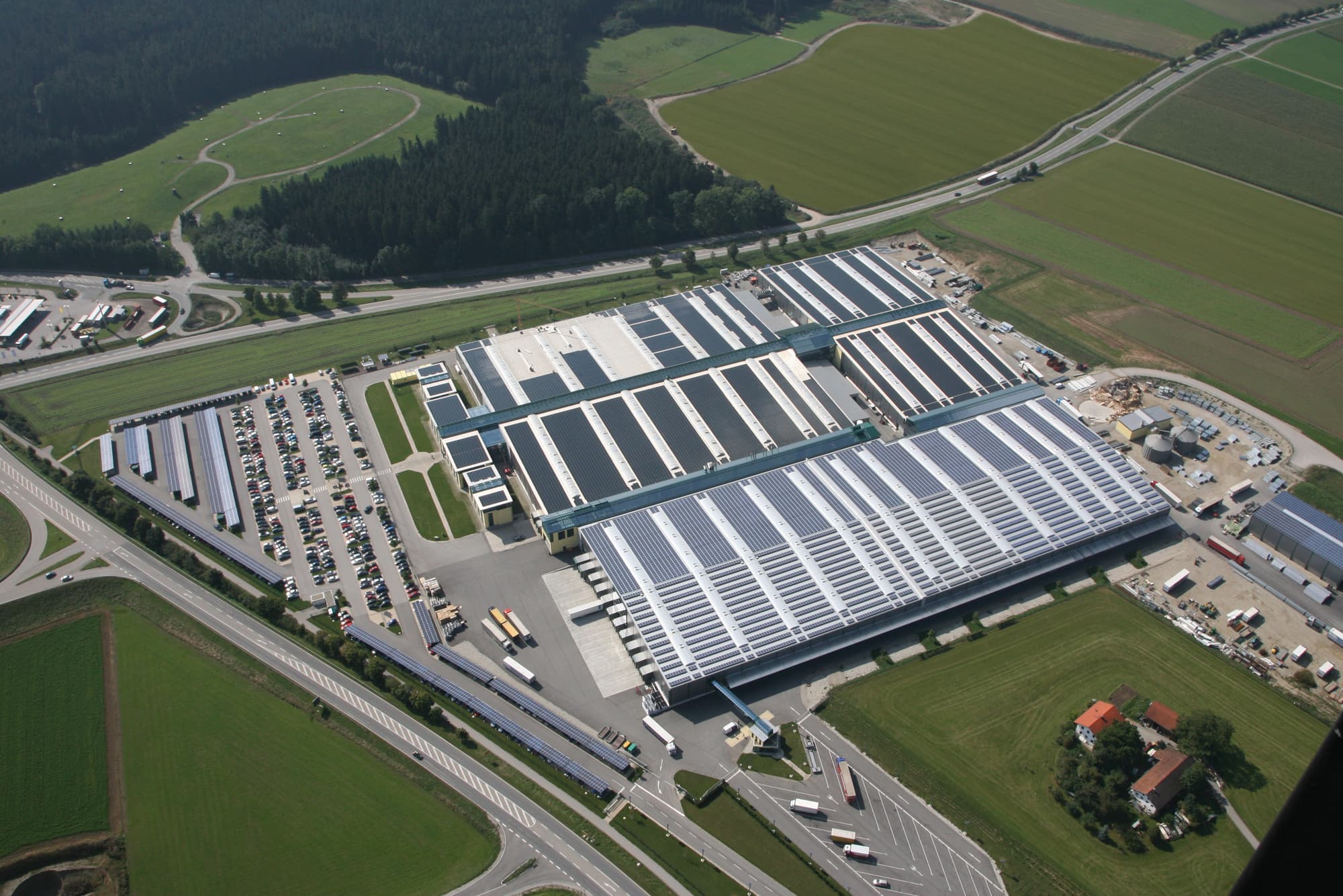 Aerial view of a large industrial facility with adjacent parking lots and surrounding green fields. Multiple buildings with white and blue rooftops are visible. Nearby roads and a small house are also shown.
