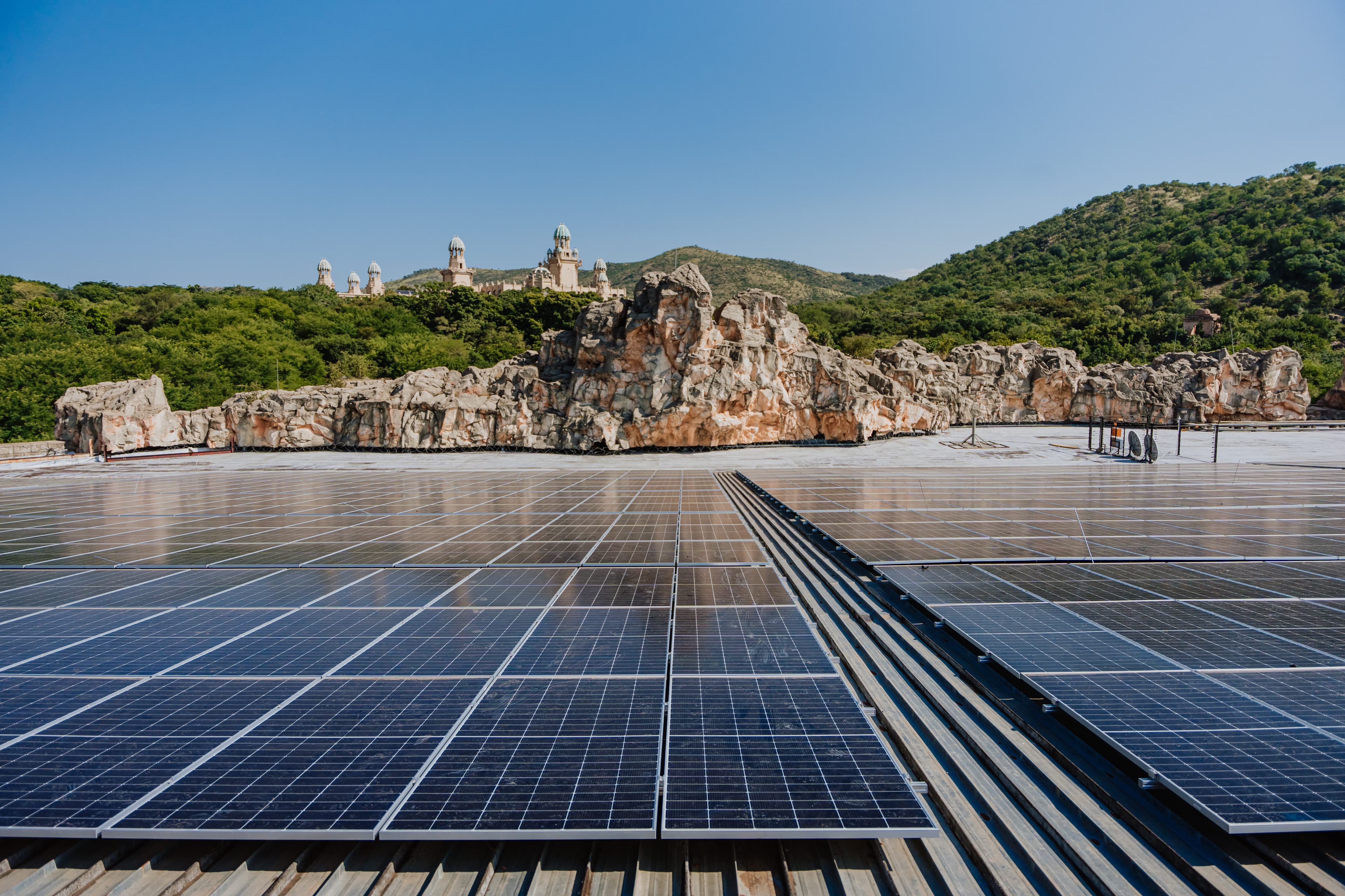 A large array of solar panels installed on a rooftop with rocky hills and a building in the background under a clear blue sky.