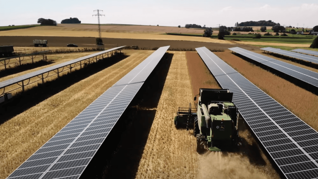 A green combine harvester operates in a field with rows of solar panels on both sides in a rural landscape.