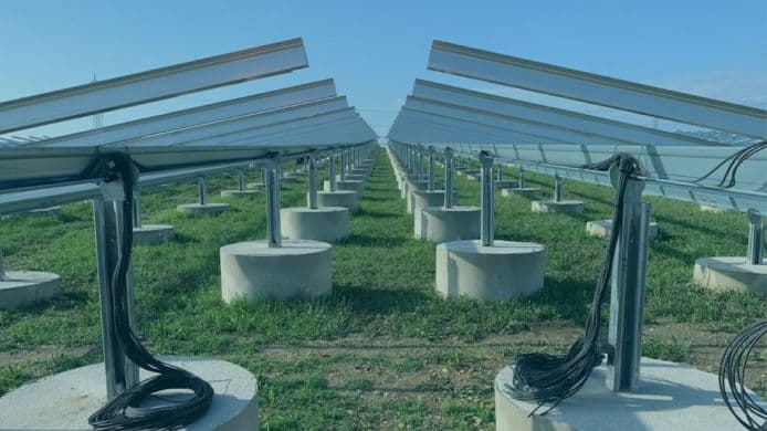 Rows of solar panel mounts in a field, ready for installation. Each mount is connected with black cables, and the ground is covered with green grass. Sky is visible in the background.