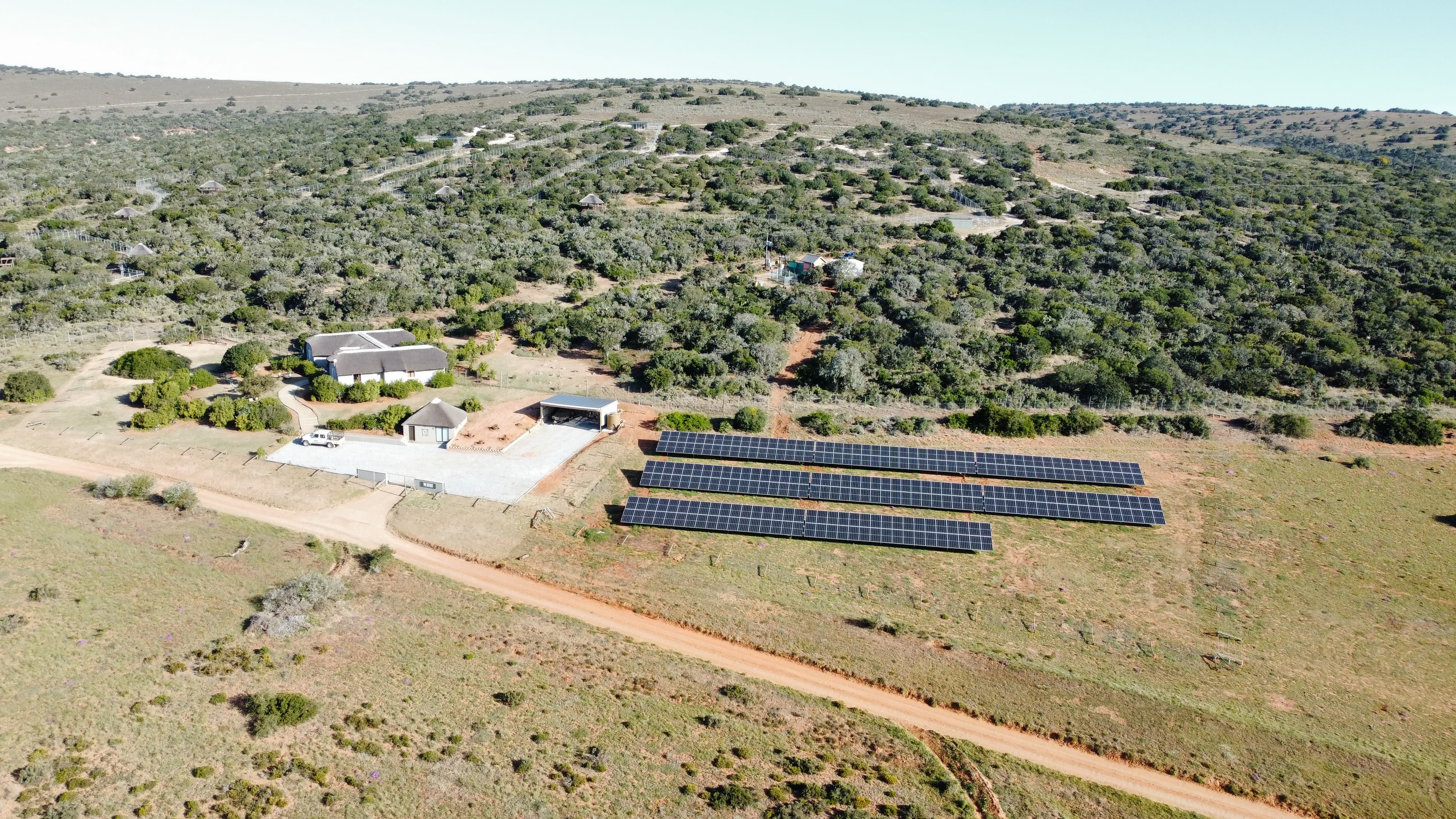 Aerial view of a rural area with solar panels installed near a building, surrounded by vegetation and dirt paths.