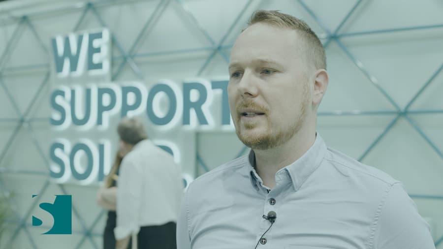 A man with short hair and beard stands in the foreground, speaking, with a sign reading "We Support Solar" and two other people in the blurred background.