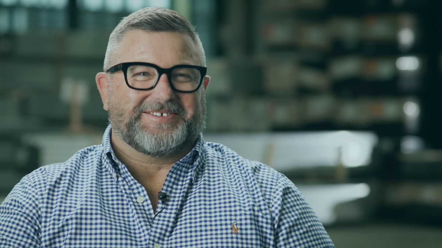 A bearded man wearing glasses and a checkered shirt smiles while seated indoors, with industrial shelving in the background.
