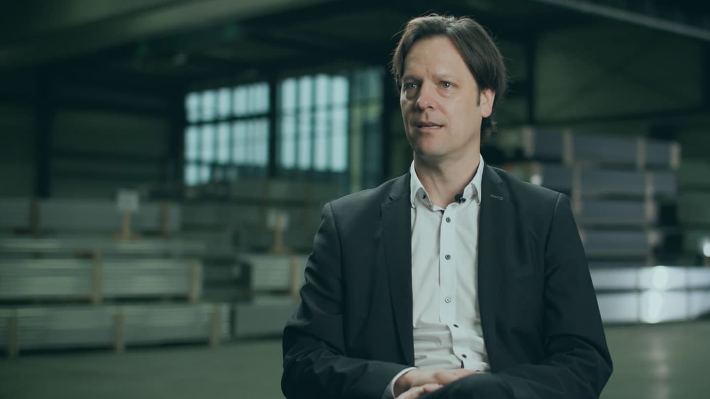 A man in a dark suit and white shirt sits in a large indoor industrial space with metal materials in the background.