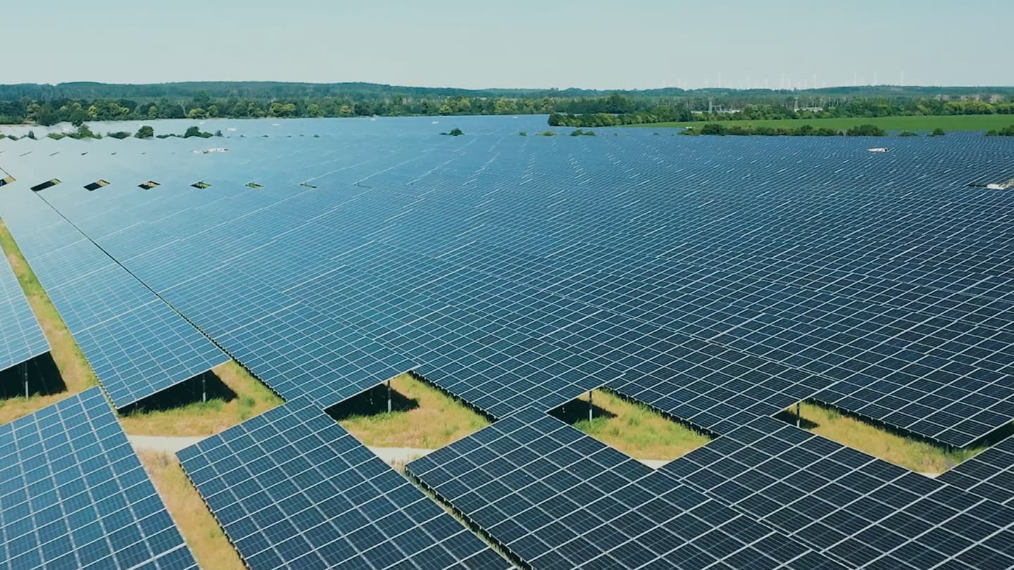 Aerial view of a large solar farm featuring numerous solar panels arranged in rows, with a landscape of trees and hills in the background under a clear sky.