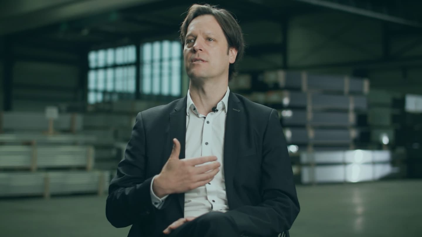 A man in a suit sits in an indoor industrial space speaking, with stacks of materials visible in the background.