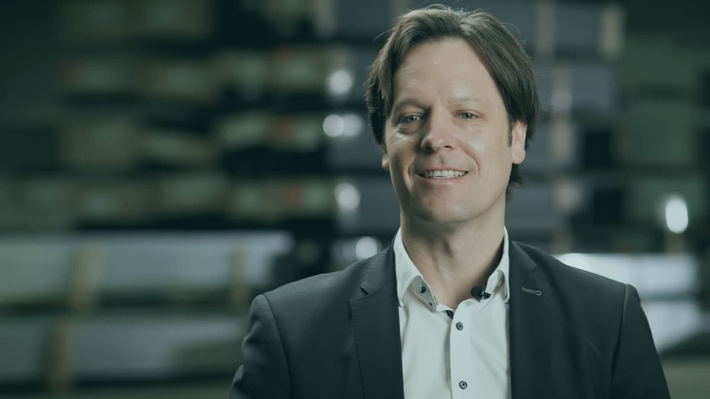 A man with short dark hair, dressed in a suit and white shirt, is smiling in front of a blurred industrial background.