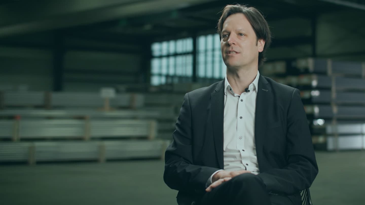 A man in a suit is seated indoors with metal shelving units in the background.