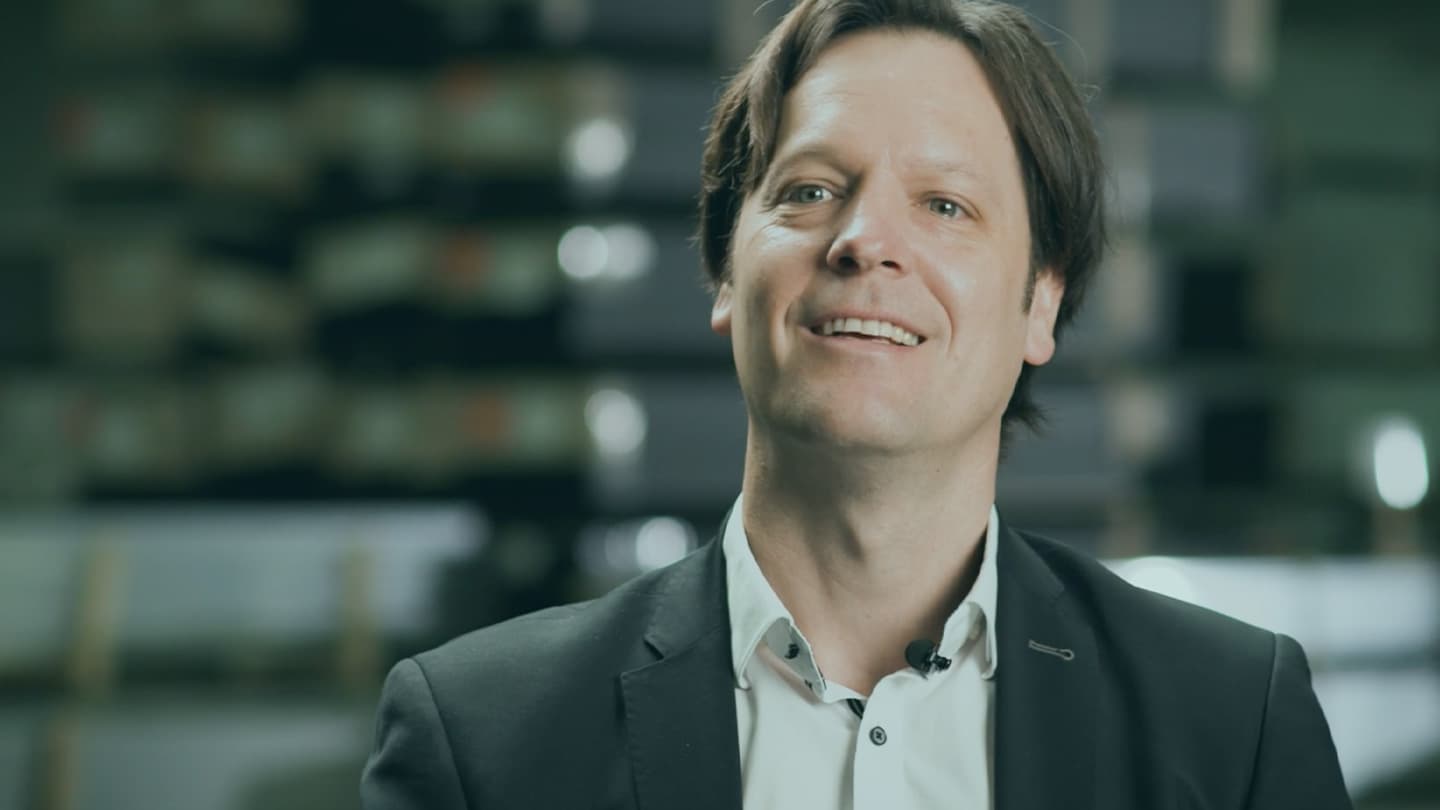 A man in a suit smiles while looking slightly off-camera. The background is blurred, with indistinct objects or shelves visible.