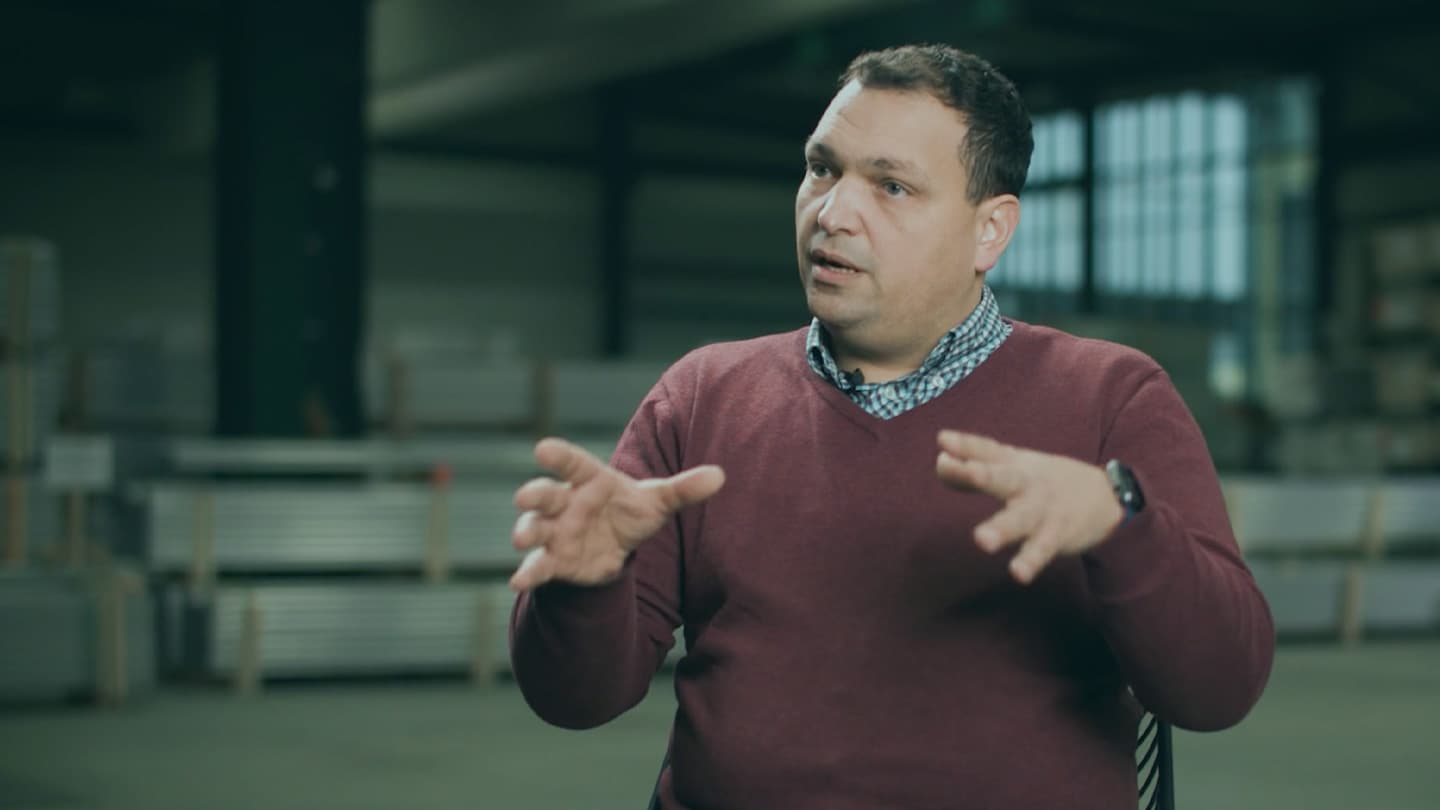 A man in a maroon sweater and blue checkered shirt is gesturing with his hands while speaking in an indoor setting.