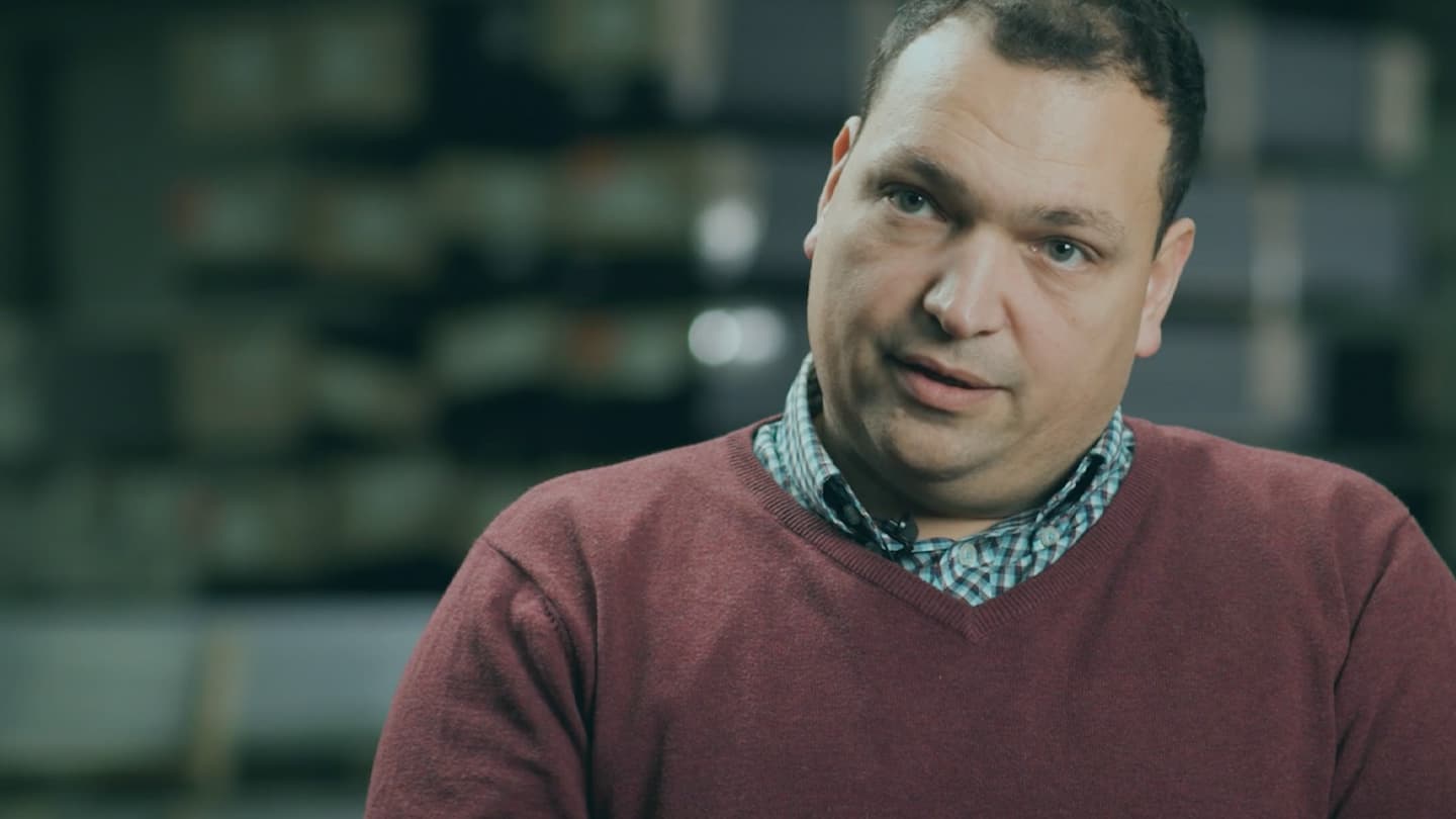 A man with short hair and a serious expression is wearing a red sweater over a checkered shirt. Shelves filled with boxes and items are blurred in the background.